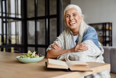 Smiling senior woman reading a book while sitting in the kitchen, eating salad